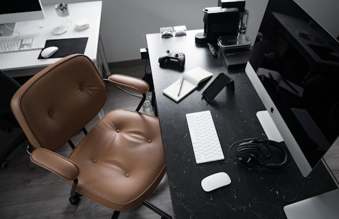 A modern workspace featuring a black desk with an Apple computer, white keyboard, and mouse. On the desk are also a brown leather chair, open notebook with pens, a smartphone on a stand, headphones, a game controller, and an espresso machine.