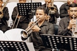 A group of musicians seated in rows, all wearing black attire, playing brass instruments like trumpets and trombones. Sheet music stands with black and white music sheets are visible in the foreground.