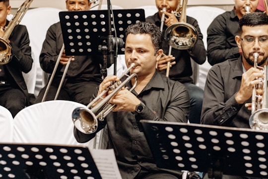 A group of musicians seated in rows, all wearing black attire, playing brass instruments like trumpets and trombones. Sheet music stands with black and white music sheets are visible in the foreground.