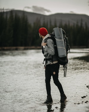 A person wearing a red beanie and gray jacket stands in a shallow body of water surrounded by a forest. They carry a large backpack equipped with camping gear, including a rolled-up sleeping mat. The background features tall evergreen trees and a range of mountains under a cloudy sky.