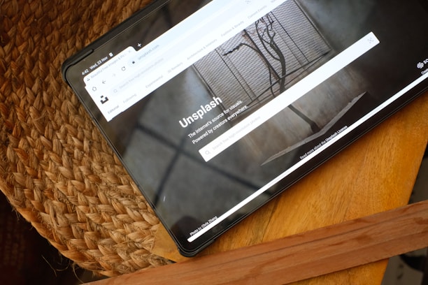 A tablet resting on a woven straw mat displays a website with a clean layout featuring a large image of a tree branch with a shadow against a concrete wall. The browser toolbar and tabs are visible, indicating an open page on Unsplash.