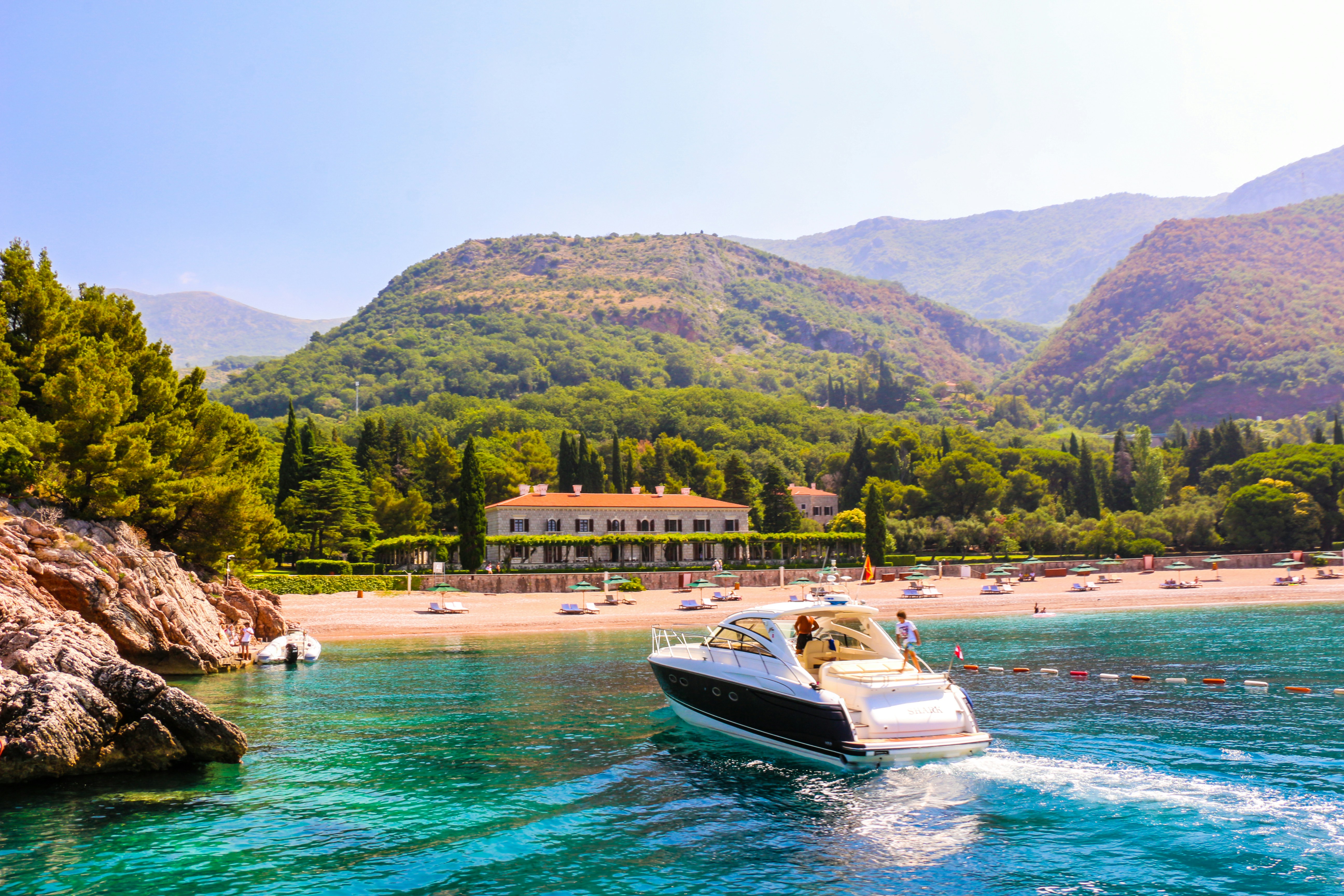A luxurious yacht sails through vibrant turquoise waters near a rocky coastline. In the background, a lush green landscape surrounds a large estate or resort, with a sandy beach dotted with sunbathers and colorful umbrellas. Rolling hills and mountains provide a picturesque backdrop, adding depth and beauty to the scene.