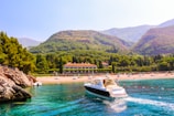 A luxurious yacht sails through vibrant turquoise waters near a rocky coastline. In the background, a lush green landscape surrounds a large estate or resort, with a sandy beach dotted with sunbathers and colorful umbrellas. Rolling hills and mountains provide a picturesque backdrop, adding depth and beauty to the scene.