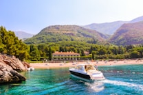 A luxurious yacht sails through vibrant turquoise waters near a rocky coastline. In the background, a lush green landscape surrounds a large estate or resort, with a sandy beach dotted with sunbathers and colorful umbrellas. Rolling hills and mountains provide a picturesque backdrop, adding depth and beauty to the scene.