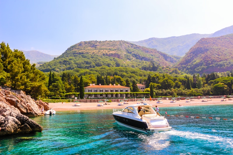 A luxurious yacht sails through vibrant turquoise waters near a rocky coastline. In the background, a lush green landscape surrounds a large estate or resort, with a sandy beach dotted with sunbathers and colorful umbrellas. Rolling hills and mountains provide a picturesque backdrop, adding depth and beauty to the scene.