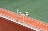 Surveillance cameras are mounted on a post beside a clay tennis court, with a green artificial turf surface visible in the background.
