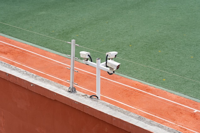 Surveillance cameras are mounted on a post beside a clay tennis court, with a green artificial turf surface visible in the background.