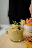 A small glass bowl filled with creamy hummus garnished with a sprig of rosemary is placed on a light wooden surface. A hand is holding a red cracker topped with sesame seeds, which has a scoop of hummus on it. Additional assorted crackers are scattered around the bowl, and there is a blurred bowl of carrot sticks in the background.