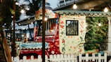 A vibrantly painted vehicle with intricate, colorful floral and geometric patterns. The windows reflect some lights, and the vehicle is parked behind a white wooden fence. The area is festively decorated with hanging string lights and surrounded by tropical plants and modern architectural elements.