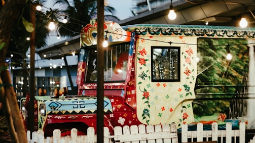 A vibrantly painted vehicle with intricate, colorful floral and geometric patterns. The windows reflect some lights, and the vehicle is parked behind a white wooden fence. The area is festively decorated with hanging string lights and surrounded by tropical plants and modern architectural elements.