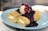A plate of two rolled crepes filled with cream cheese, topped with a scoop of vanilla ice cream and drizzled with a rich berry sauce, garnished with whole blueberries.