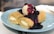 A plate of two rolled crepes filled with cream cheese, topped with a scoop of vanilla ice cream and drizzled with a rich berry sauce, garnished with whole blueberries.