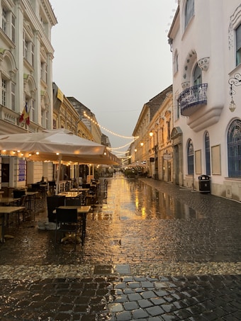 A picturesque cobblestone street glistens with rain, bordered by elegant historical buildings with ornate architectural features. Warm string lights hang overhead, casting a cozy glow on outdoor seating areas sheltered by awnings. The ambiance suggests a quiet, serene evening in a charming European town.