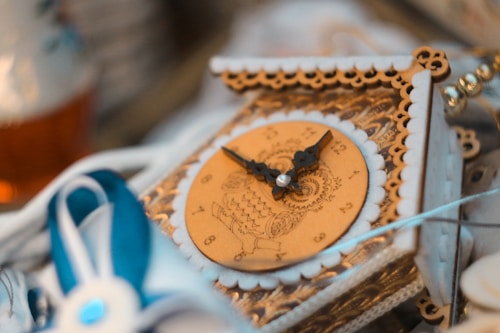 An intricately designed ornamental wooden clock with delicate cutouts and detailed engravings, featuring ornate clock hands and a whimsical number layout. Accompanying the clock are elements of lace and decorative blue ribbons.