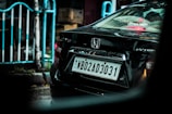 A black car is parked on a wet street, displaying a license plate with the code WB02AD3031. The back of the car is shown, featuring a Honda logo and model markings. Reflections and raindrops are visible on the car's surface. A white and blue railing and blurred city elements are in the background.