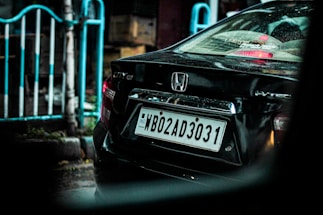 A black car is parked on a wet street, displaying a license plate with the code WB02AD3031. The back of the car is shown, featuring a Honda logo and model markings. Reflections and raindrops are visible on the car's surface. A white and blue railing and blurred city elements are in the background.