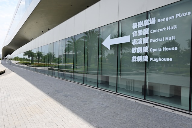 A modern building with a sleek, curved glass exterior. Large windows reflect the surrounding greenery and palm trees. White text on the glass provides directions to various cultural venues like a concert hall, recital hall, opera house, and playhouse.