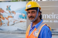 A construction worker wearing a yellow safety helmet and an orange safety vest is standing in front of a billboard featuring an image of people lounging on a yacht. The worker has a slight smile and is looking at the camera.