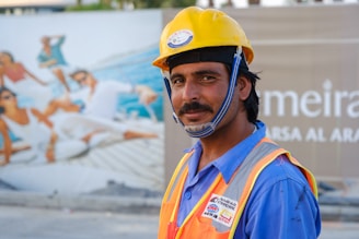 A construction worker wearing a yellow safety helmet and an orange safety vest is standing in front of a billboard featuring an image of people lounging on a yacht. The worker has a slight smile and is looking at the camera.