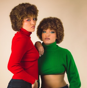Two individuals with similar hairstyles are wearing vibrant knit turtlenecks, one in red and the other in green, against a neutral background. They are posed closely together, with one person resting an arm on the other's shoulder.