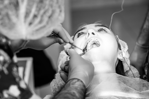 A close-up black and white image of a dental procedure in progress. A patient is lying back with their mouth open, while a dentist or dental assistant, wearing gloves and a hair covering, is working on the patient's teeth using dental instruments. The patient appears to be wearing a dental bib and some medical equipment is visible in the background.