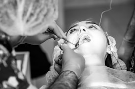 A close-up black and white image of a dental procedure in progress. A patient is lying back with their mouth open, while a dentist or dental assistant, wearing gloves and a hair covering, is working on the patient's teeth using dental instruments. The patient appears to be wearing a dental bib and some medical equipment is visible in the background.