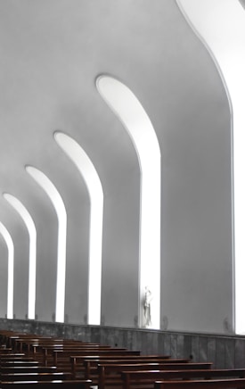 A modern architectural interior featuring tall, narrow, arched windows that let in soft light, creating a serene and peaceful atmosphere. Wooden pews are arranged in rows, suggesting a place of worship or meditation. A statue stands by one of the windows, adding a spiritual element to the space.