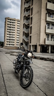 A motorbike is parked on a concrete surface in front of a tall residential building with multiple balconies. There is another high-rise building in the background. The sky is overcast, contributing to a somber atmosphere. The motorbike's headlight is on, and there are puddles on the ground indicating recent rainfall.