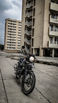 A motorbike is parked on a concrete surface in front of a tall residential building with multiple balconies. There is another high-rise building in the background. The sky is overcast, contributing to a somber atmosphere. The motorbike's headlight is on, and there are puddles on the ground indicating recent rainfall.