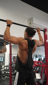 A person is performing a pull-up exercise in a gym setting. They are wearing a black sleeveless shirt, black and white track pants, and a watch. The gym is equipped with dumbbells, mirrors, and red gym equipment in the background.