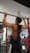A person is performing a pull-up exercise in a gym setting. They are wearing a black sleeveless shirt, black and white track pants, and a watch. The gym is equipped with dumbbells, mirrors, and red gym equipment in the background.