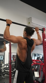 A person is performing a pull-up exercise in a gym setting. They are wearing a black sleeveless shirt, black and white track pants, and a watch. The gym is equipped with dumbbells, mirrors, and red gym equipment in the background.