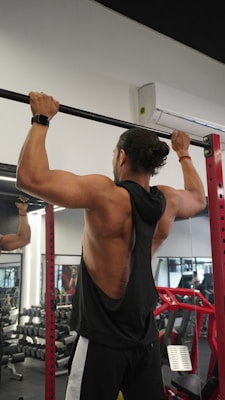 A person is performing a pull-up exercise in a gym setting. They are wearing a black sleeveless shirt, black and white track pants, and a watch. The gym is equipped with dumbbells, mirrors, and red gym equipment in the background.