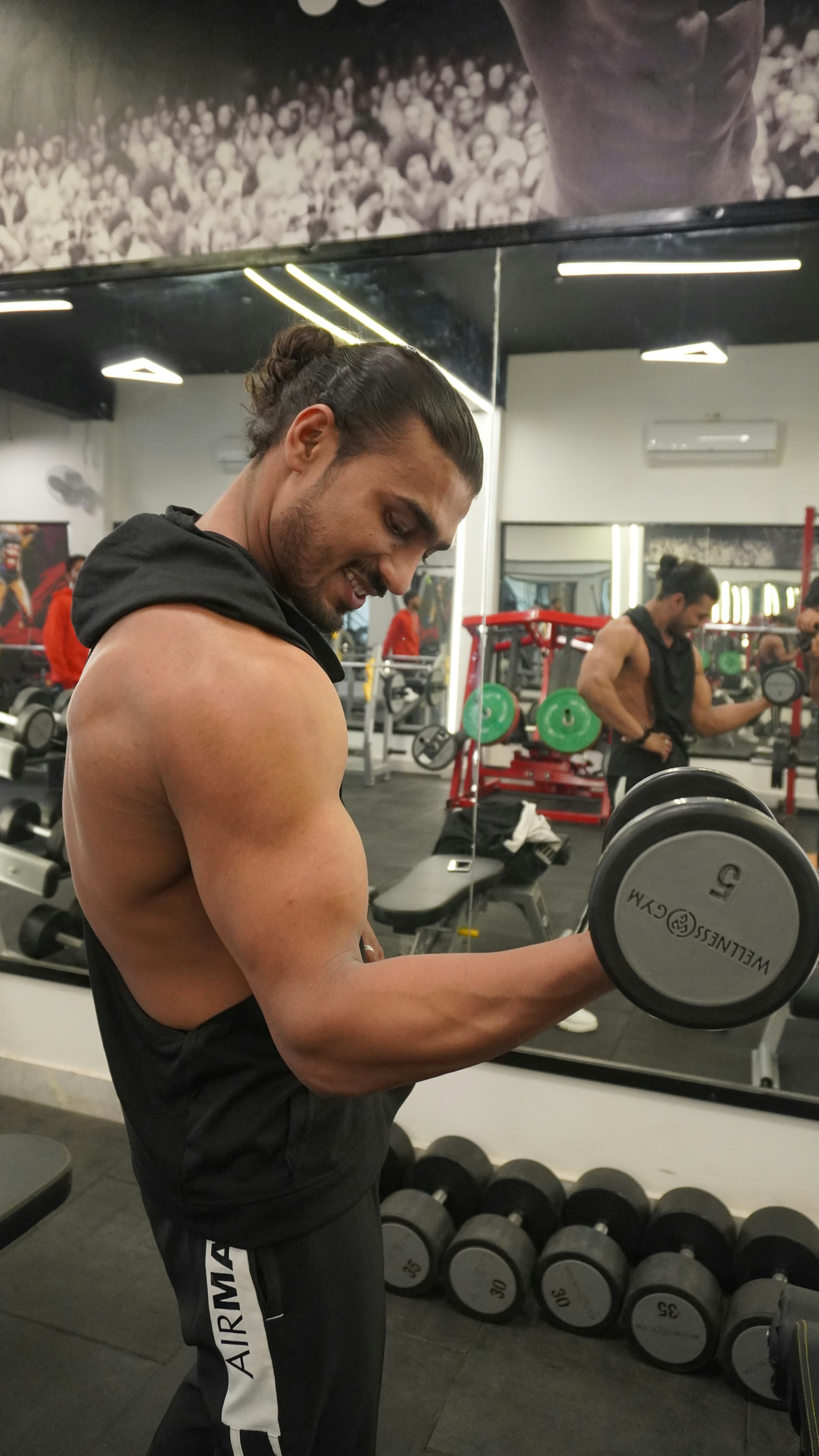 A muscular person lifts a dumbbell in a gym, standing in front of a mirror with various gym equipment visible in the background. The individual has long hair tied back and is wearing a sleeveless top.