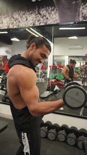 A muscular person lifts a dumbbell in a gym, standing in front of a mirror with various gym equipment visible in the background. The individual has long hair tied back and is wearing a sleeveless top.