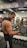 A muscular person lifts a dumbbell in a gym, standing in front of a mirror with various gym equipment visible in the background. The individual has long hair tied back and is wearing a sleeveless top.