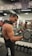 A muscular person lifts a dumbbell in a gym, standing in front of a mirror with various gym equipment visible in the background. The individual has long hair tied back and is wearing a sleeveless top.
