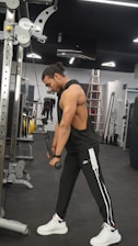 A man with long hair tied back is working out in a gym, using a cable machine to perform a tricep pushdown exercise. He is dressed in a sleeveless black top and black track pants with white and black stripes, wearing white sneakers. The gym has modern equipment and a sleek interior, with a ladder visible in the background.