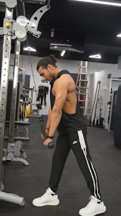 A man with long hair tied back is working out in a gym, using a cable machine to perform a tricep pushdown exercise. He is dressed in a sleeveless black top and black track pants with white and black stripes, wearing white sneakers. The gym has modern equipment and a sleek interior, with a ladder visible in the background.
