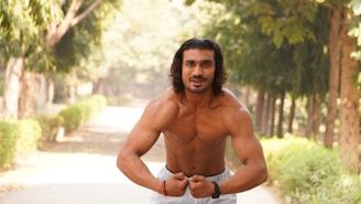 A muscular man is posing shirtless in a wooded outdoor area, flexing his biceps with a confident expression. Trees and greenery line the path, creating a peaceful, natural setting.