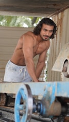 A shirtless man with long hair and a muscular physique is leaning over a piece of machinery in an industrial setting. He is wearing light-colored shorts and appears focused.
