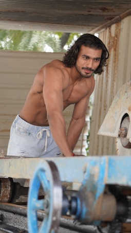 A shirtless man with long hair and a muscular physique is leaning over a piece of machinery in an industrial setting. He is wearing light-colored shorts and appears focused.