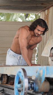 A shirtless man is working near a large industrial machine, surrounded by a rustic setting. The machinery appears to be heavy-duty with gears and a cutting component. Background elements indicate an outdoor environment with some greenery.