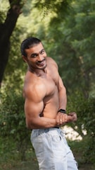 A muscular man with a friendly smile poses outdoors with greenery in the background. He is shirtless, wearing light denim jeans, and showing off his physique with a confident stance. The natural lighting highlights his toned muscles and features.