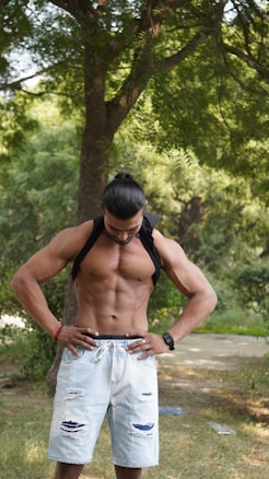 A muscular person with long dark hair is standing outdoors in a park-like setting, looking downwards. They are wearing light blue ripped denim shorts and have a black garment hanging from their neck. The background features lush green trees and a sunlit path, suggesting a serene and natural environment.
