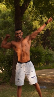 A shirtless man is standing outdoors, flexing his muscles while smiling confidently. He is wearing light-colored, ripped denim shorts and posing under a tree with lush green foliage in the background.