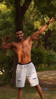 A shirtless man is standing outdoors, flexing his muscles while smiling confidently. He is wearing light-colored, ripped denim shorts and posing under a tree with lush green foliage in the background.