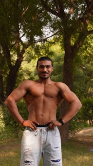 A muscular, shirtless man with his hands on his hips stands confidently in a wooded area. Tall trees surround him, creating a natural and serene backdrop. He is wearing ripped light blue jeans and a watch on his left wrist.