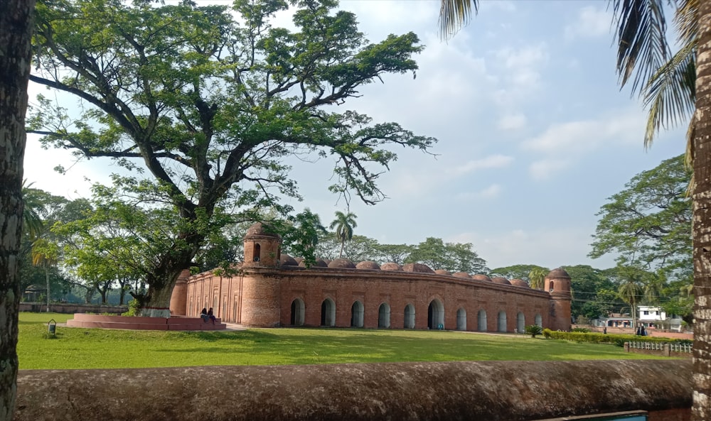 a large brick building with a lawn and trees in front of it with Sixty Dome Mosque in the background