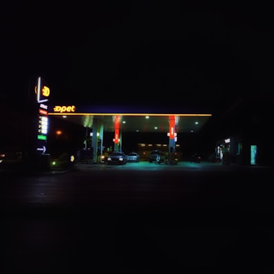 A petrol station is illuminated at night, featuring a bright canopy with red and yellow branding. Several cars are parked under the shelter, and the surrounding area is enveloped in darkness, highlighting the contrast with the well-lit petrol station.